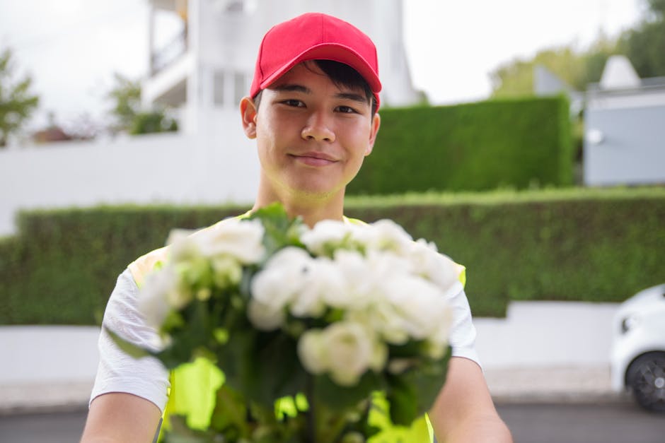 A delivery person, wearing a red cap and a high-visibility vest, extends a bouquet of fresh white flowers towards the camera, smiling gently. The bouquet features a variety of white blooms, possibly r