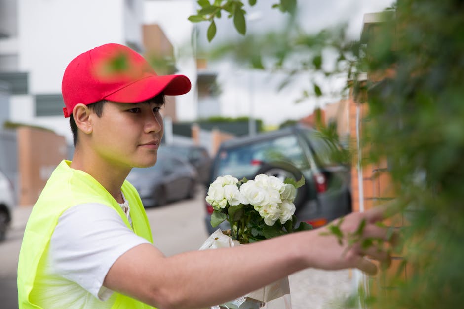 A young delivery person wearing a red cap and a neon yellow vest is shown reaching out to place a bouquet of fresh white flowers, possibly hydrangeas, wrapped in white paper, against a green shrub out