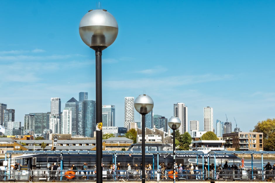 A cityscape with a clear blue sky featuring modern skyscrapers in the background and a riverside walkway in the foreground. The walkway is lined with tall, spherical street lamps, and a glass-enclosed