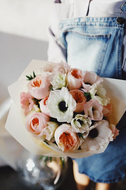 A person dressed in a light-colored top and blue jeans is holding a bouquet of fresh flowers wrapped in cream-colored paper. The bouquet features a mix of pale pink peonies, peach roses, and white ane