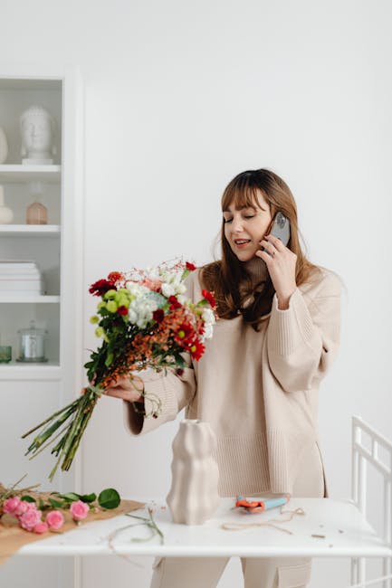 A woman in a cream-colored sweater holds a vibrant floral bouquet consisting of red, white, pink, and green flowers, including roses and daisies, with visible fresh petals and green stems. She is spea