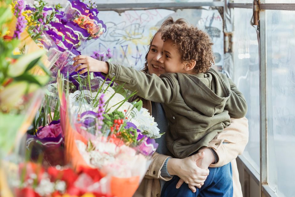 A young boy with curly hair and a smile, being held by an adult, is reaching out to select a colorful arrangement of fresh flowers at a flower shop. The bouquet features a variety of vibrant blooms in
