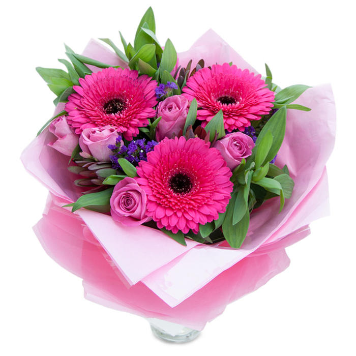 I imagine setting this bouquet down on a kitchen island near the Thames: three exuberant fuchsia Gerbera daisies open wide, their long petals forming a radiant halo around deep, almost black centres that lend bold contrast and visual rhythm. Clustered close, lavender-pink roses in a dusty blush hue curl inward, their tightly furled petals catching light in soft, velvety bands that invite a gentle touch. Scattered throughout are small, vivid clusters of purple statice, their papery texture providing a playful punctuation that breaks the pink palette in the most pleasing way. A generous sweep of green foliage-leaves that read as salal and eucalyptus-wraps the composition, their cool green tones and subtle scent grounding the bouquet with a fresh, botanical voice. The arrangement is lovingly wrapped: a matte pale pink tissue acts as a soft, protective base and is overlaid by a slightly crinkled, translucent fuchsia-pink cellophane that adds an airy shimmer and a polished finish. Peeking from beneath is the top rim of a clear glass vase, hinting that these blooms are meant to be displayed immediately and enjoyed up close. In dawn or late-afternoon light the roses give off a gentle perfume, the gerberas' subtle green sweetness balances the floral notes, and the statice holds its colour like a tiny chorus of accents. Sized and styled to suit celebrations across Blackwall, near the Blackwall Tunnel approach or in an apartment overlooking Canary Wharf, this bouquet offers a mix of playfulness and grace that feels distinctly local and thoughtfully made.