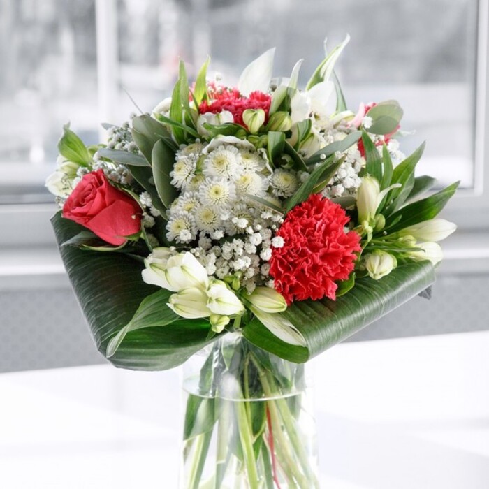 A refined arrangement of red and white flowers takes centre stage in a clear cylindrical glass vase, the kind you might find on a neatly set table in a Blackwall townhouse overlooking the river. The vase is filled with fresh water, revealing a bundle of carefully trimmed green stems, lined up straight and clean to suggest both freshness and professional care. At the heart of the bouquet, rich deep red roses unfurl softly, each velvety petal catching the light and adding a sense of classic romance. Beside them sits a large crimson carnation, its petals tightly layered and intricately ruffled, bringing a tactile, almost sculptural quality to the composition. Interwoven with these vivid reds are white flowers that create a tender contrast: alstroemeria with slim, lightly speckled petals, creamy chrysanthemum-like blooms that feel full and generous, and several white lily buds that remain poised and closed, suggesting beauty still to come. Fine sprays of gypsophila, or baby's breath, float between the larger blooms like a gentle mist, making the overall design feel airy and abundant. The arrangement is framed with lush greenery: broad, glossy dark green leaves are skillfully wrapped and curled around the base to form a polished collar, while varied foliage in eucalyptus-like shapes and pointed leaves adds layers of texture and shifting shades of green. The bouquet rests on a crisp white surface against a bright, out-of-focus background, as if placed near a window looking toward the paths and parks around Blackwall. Soft, even lighting highlights every curve and edge, giving the impression of a bouquet designed as a true centrepiece-equally suited to a romantic dinner, a heartfelt thank-you, or a sophisticated gift for a home or office in this corner of East London.